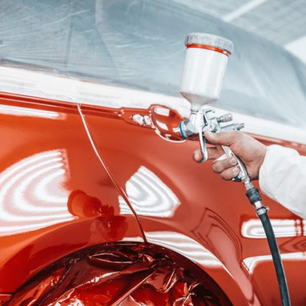 Professional painting process: A person in protective gear sprays a glossy red car with paint in a workshop using a professional spray gun.