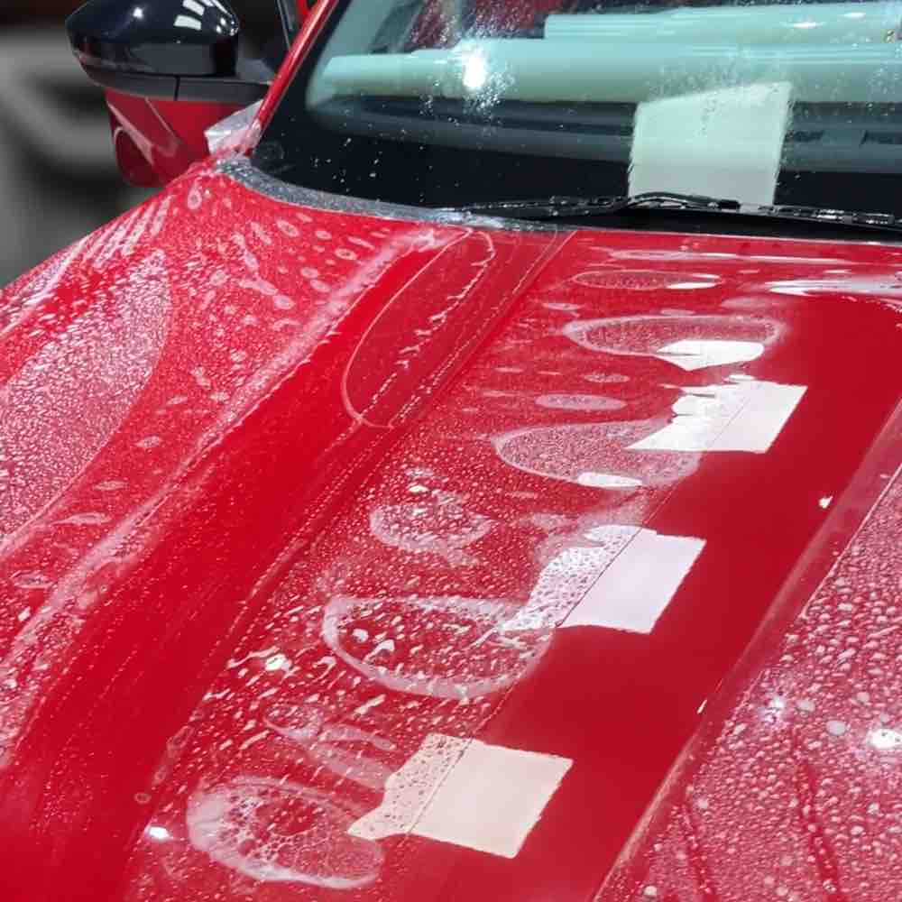 Close-up view of a partially cleaned red car with soap foam during paint protection film application. Shiny surface contrasts with foam textures, featuring PPF Tool 'Squeegee.'
