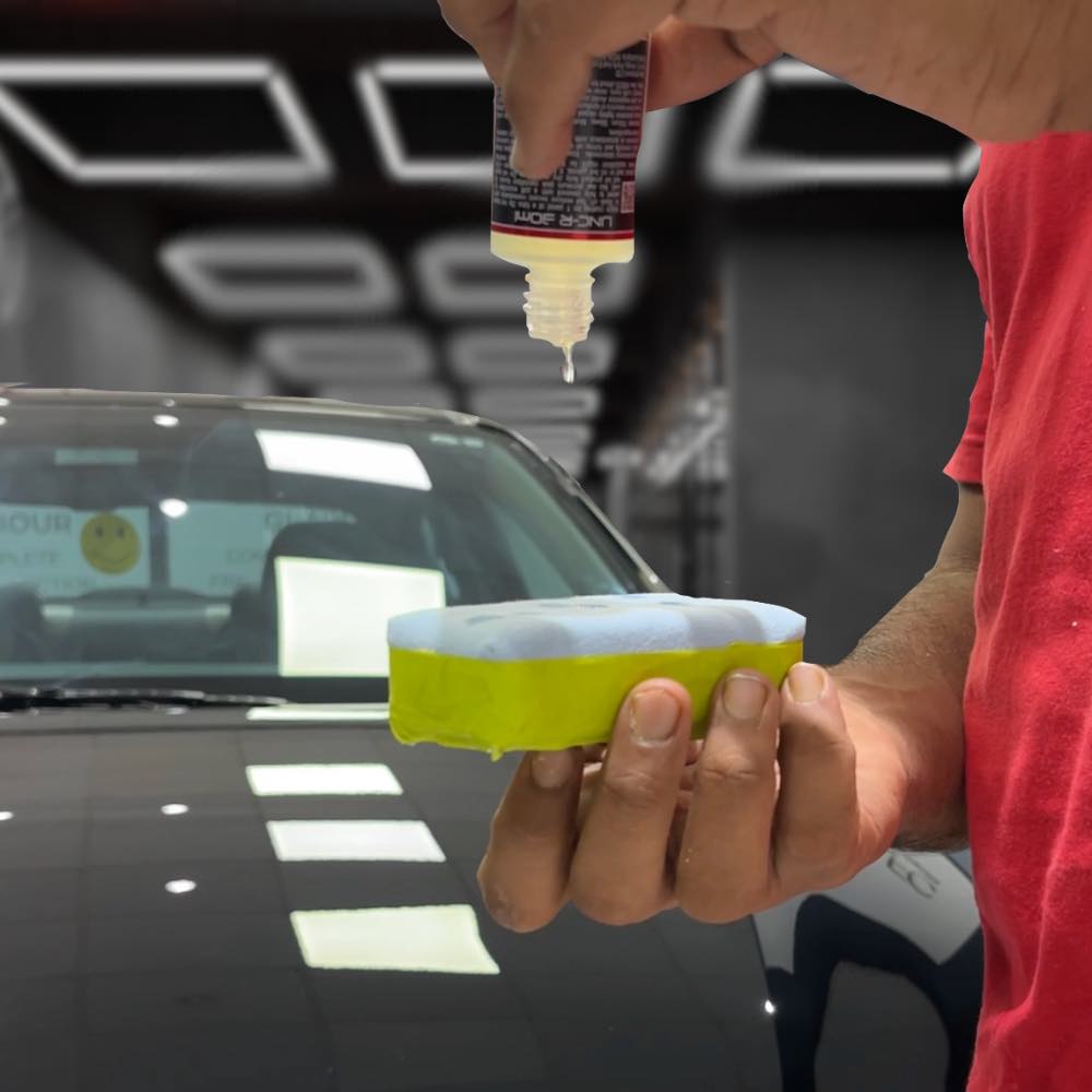 Person applying Ceramic Coating liquid from a small bottle onto a sponge for car detailing and paint protection. Black car in an indoor workshop setting.