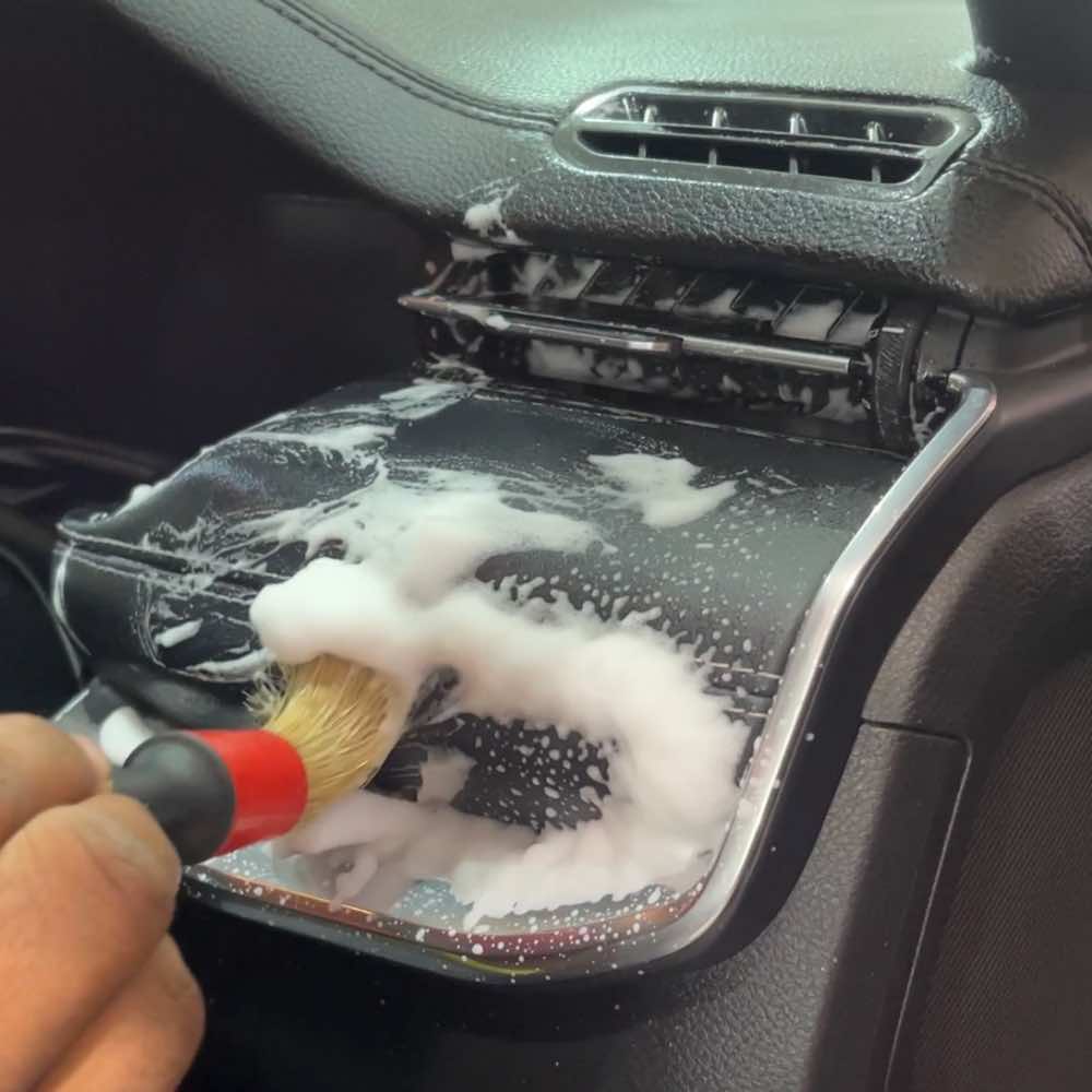 Car interior cleaning: Brush and foam used to clean air vent and surrounding area. Partially disassembled vent reveals internal structure for thorough cleaning of the black dashboard.